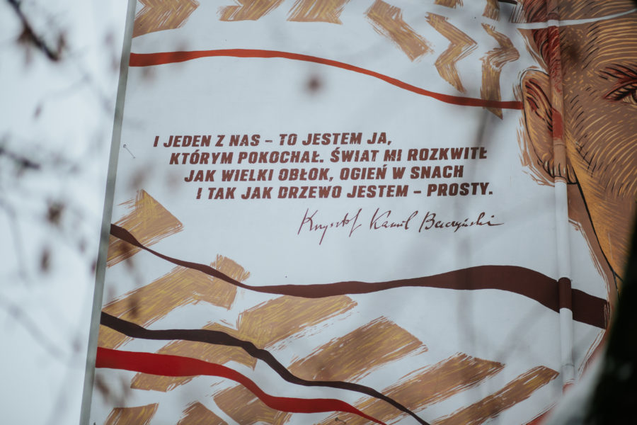 longdesc= "Zbliżenie na cytat znajdujący się na muralu, który brzmi: jeden z nas — to jestem ja,. którym pokochał. Świat mi rozkwitł. jak wielki obłok, ogień w snach. i tak jak drzewo jestem — prosty."