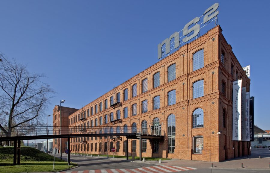 Industrialny budynek z czerwonej cegły, na jego dachu przymocowane są wielkie metalowe znaki: „MS do kwadratu”.