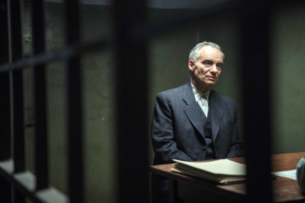 Fotos z filmu „Szarlatan”. Za kratami przy stole rozmawia elegancko ubrany mężczyzna z kimś nieujętym w kadrze. Na brzegu stołu leży plik papierów.