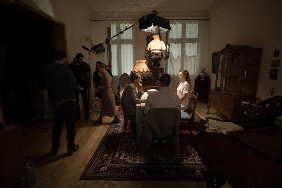 Kulisy powstawania filmu „Położna z Auschwitz”. Trzech dorosłych siedzi przy stole. Nad nimi znajduje się lampa filmowa. Z lewej strony stoi kilku członków ekipy filmowej.