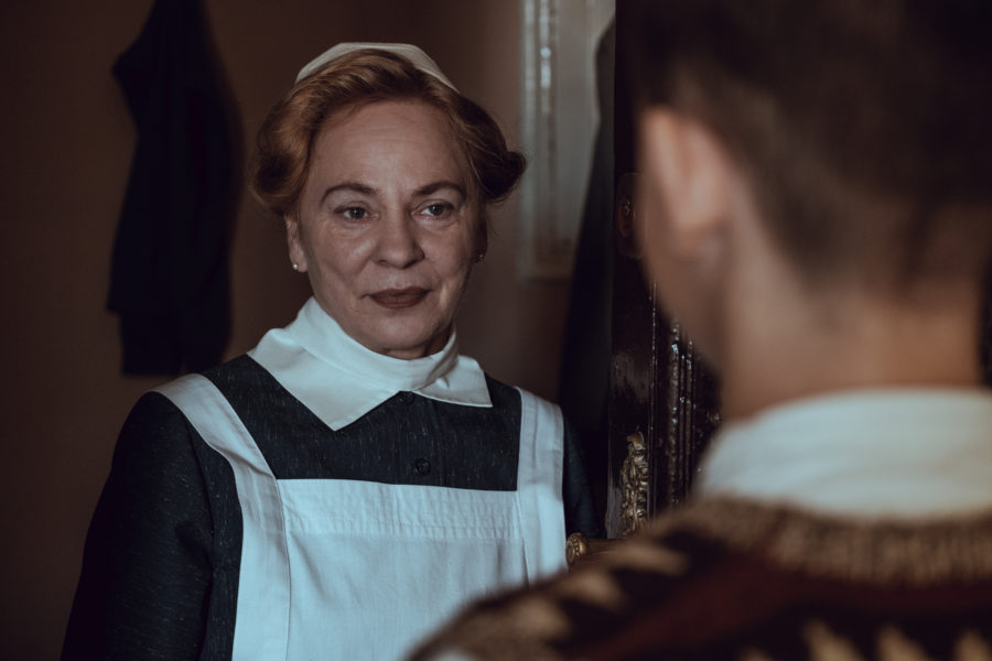 Kadr z filmu „Położna z Auschwitz”. Starsza kobieta w fartuchu otwiera drzwi wejściowe młodemu mężczyźnie.
