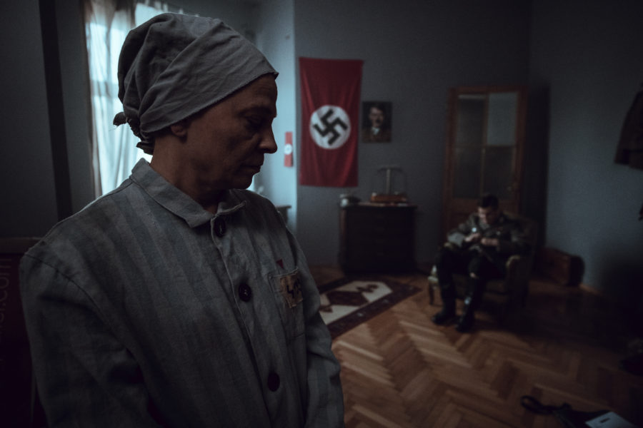 Kadr z filmu. Gabinet oficera Trzeciej Rzeszy. Mężczyzna w mundurze siedzi na krześle. Na pierwszym planie stoi starsza kobieta w więziennym pasiaku.