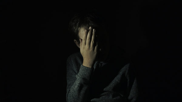 Częściowo oświetlony chłopczyk, zasłania ręką część twarzy. Chłopczyk znajduje się na ciemnym tle.