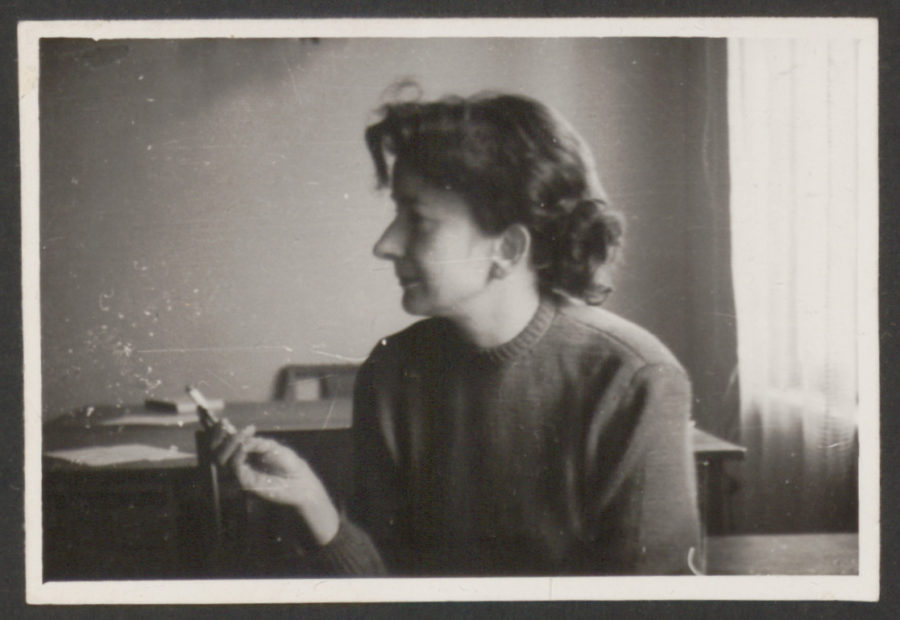 Młoda Wiesława Szymborska siedzi obrócona profilem. W ręce trzyma papierosa.