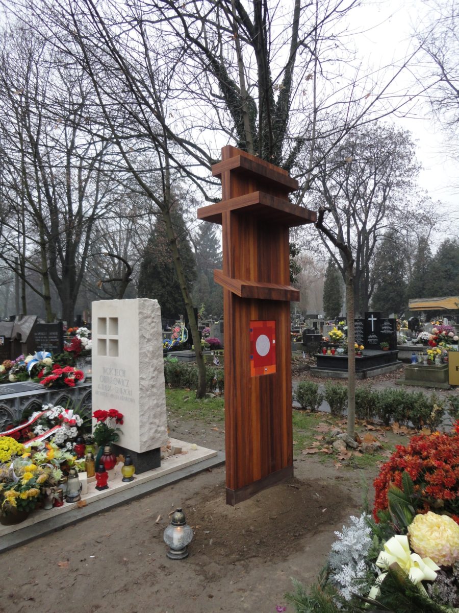 Drewniany, wysoki krzyż prawosławny stoi wbity w ziemię. Przed nim znajduje się jeden znicz.