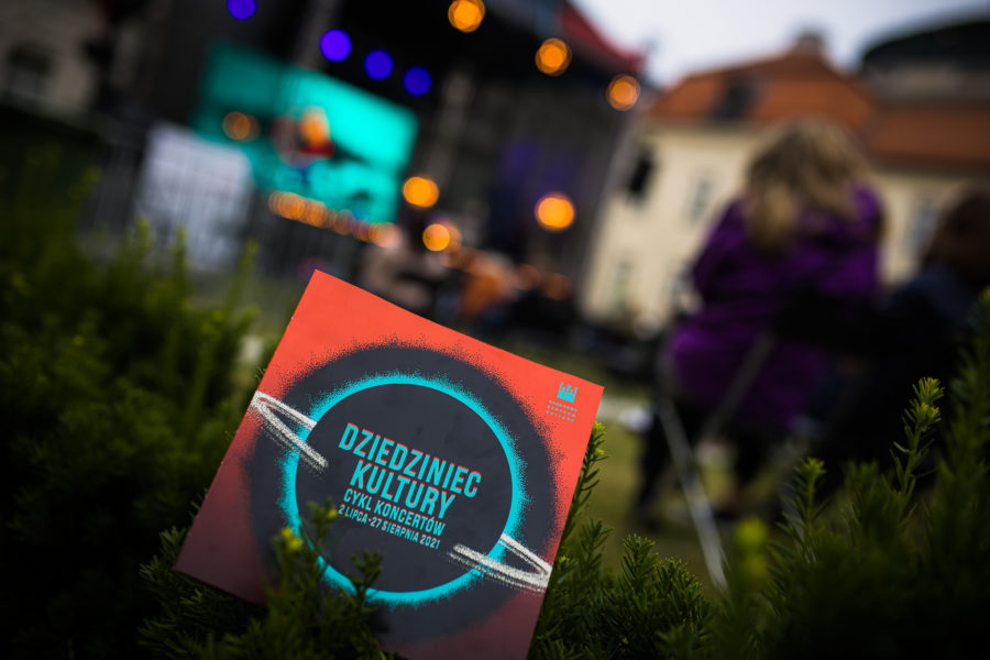 Wlepka z napisem: "Dziedziniec Kultury" leży na trawie na tle sceny.
